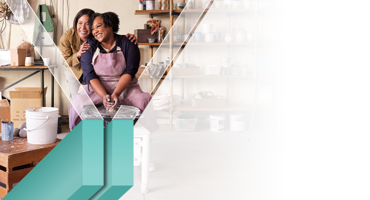 A woman happily makes pottery as her friend stands behind her giving an encouraging hug.